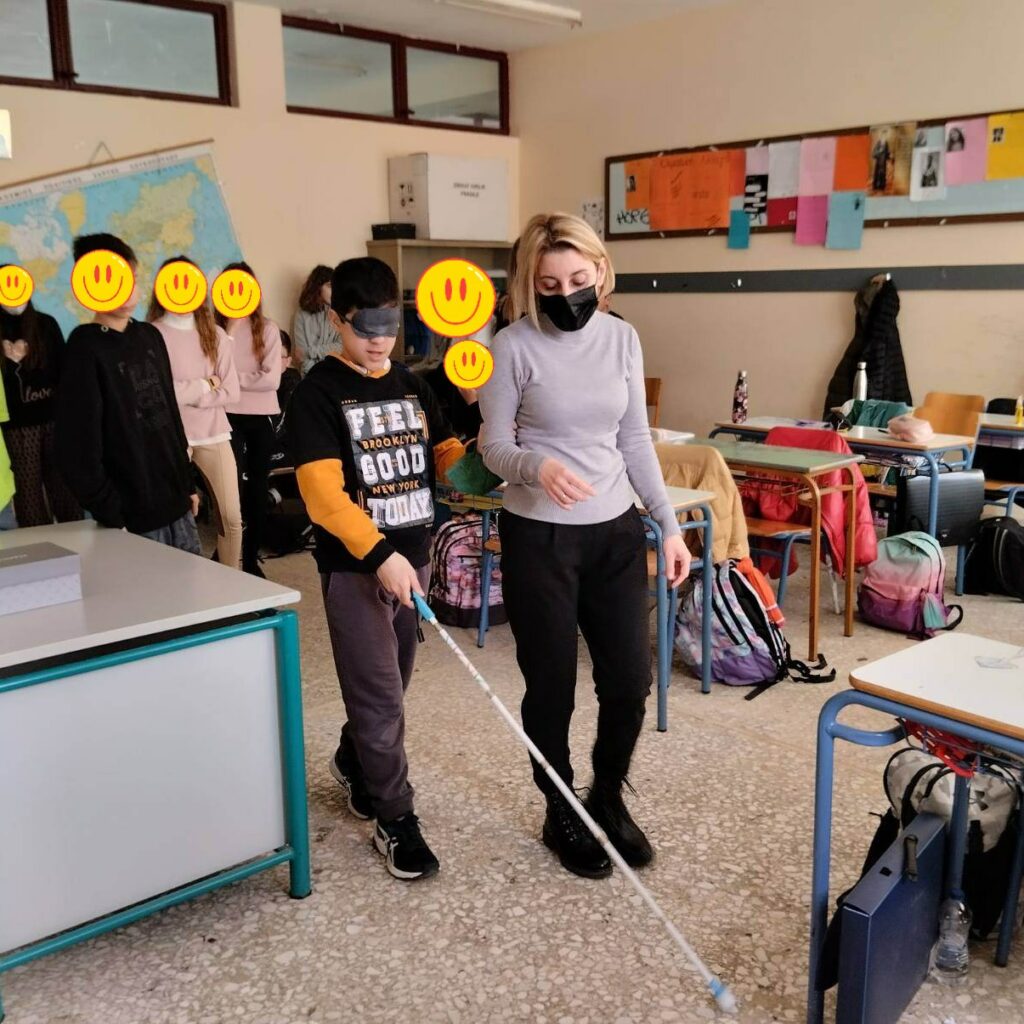 Η εκπαιδεύτρια Κινητικότητας, Προσανατολισμού και Δεξιοτήτων Καθημερινής Διαβίωσης, Γεωργία Κυριτσοπούλου, βοηθάει μαθητή που καλύπτει τα μάτια του με μάσκα πως να χειρίζεται το λευκό μπαστούνι. 