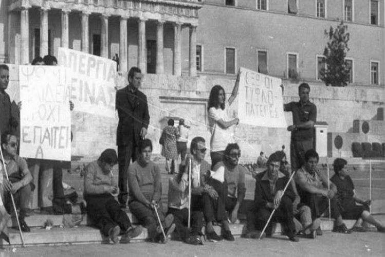 Στιγμιοτυπο από τη διαμαρτυρία των τυφλών με πανό έξω από τη Βουλή των Ελλήνων. Κάποιοι είναι καθιστοί με λευκά μπαστούνια στο πεζοδρόμιο και κάποιοι όρθιοι κρατούν πανό με συνθήματα.