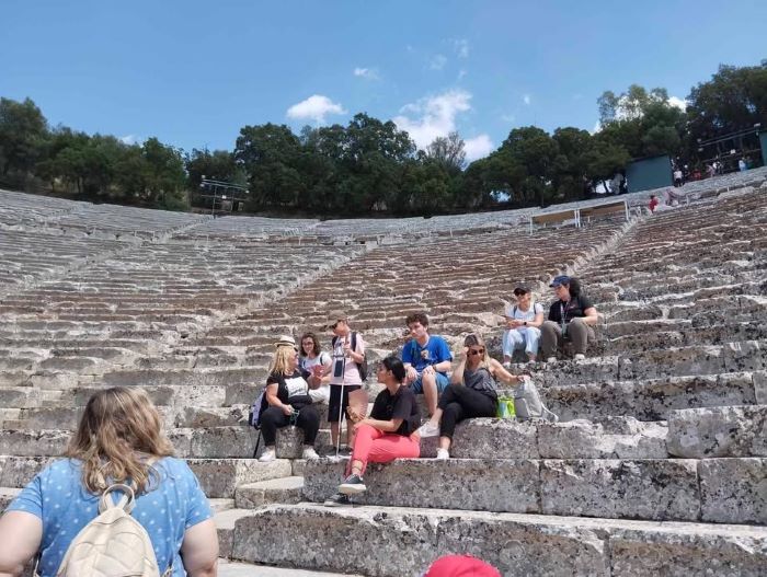 Τα παιδιά σκορπισμένα στις κερκίδες του αρχαίου θεάτρου παρακολουθούν με ενδιαφέρον τα όσα τους λέει η ξεναγός