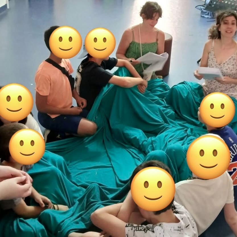 ωφελούμενοι καθισμένοι στο πάτωμα σε κύκλο παρακολουθούν το παραμύθι “ Το Αίνιγμα του Μαγεμένου Δάσους”. Είναι καλυμμένοι με πράσινο κάλυμμα.